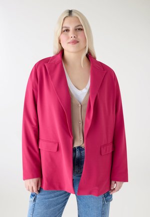 Giovane donna con lunghi capelli biondi che indossa un blazer rosa acceso, un cardigan beige con bottoni, una t-shirt bianca e jeans blu su sfondo bianco.