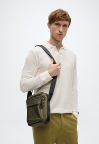 Πράσινο νάιλον τσαντάκι ώμου με φερμουάρ στην μπροστινή τσέπη και ρυθμιζόμενο μαύρο λουρί, που κρατιέται από ένα άτομο με ανοιχτόχρωμο μακρυμάνικο πουκάμισο.