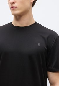 Svart t-shirt med kort ärm i texturerat tyg, med rund halsringning och en liten tryckt accentlogotyp på vänster bröst.