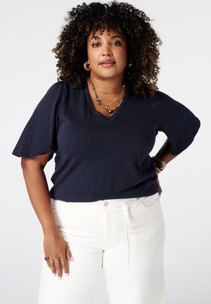 Femme aux cheveux bouclés portant un haut bleu marine à manches courtes et un pantalon blanc, debout avec une main sur la hanche et une expression neutre.