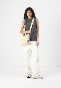 Borsa in tela beige con due manici in alto e una tracolla, dotata di una tasca anteriore e di una patch con logo, abbinata a una canotta scura e pantaloni bianchi.