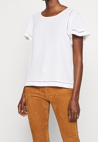 Haut blanc à manches courtes avec un col rond, détails découpés élaborés le long des manches et de l'ourlet, associé à un pantalon en daim marron.