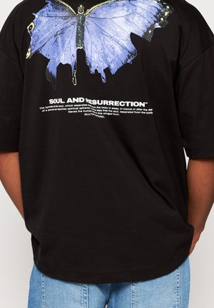 Czarny t-shirt z dużym niebieskim motylem i białym napisem "Soul and Resurrection", noszony z jasnoniebieskimi dżinsami.