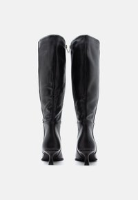 Bottes noires montant jusqu'aux genoux en cuir lisse. Dotées d'un talon étroit et d'un détail zippé à l'arrière. Design élégant et ajusté qui sublime la silhouette.
