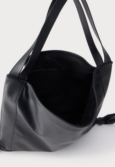 Sac bandoulière en cuir noir avec larges lanières, ouverture supérieure non fermée par zip et un nœud lâche sur un côté sur fond blanc uni.