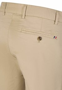 Beige Hosen aus glattem Stoff, mit einer Gesäßtasche mit Knopf und einem kleinen dreifarbigen Akzent. Dezente Nahtdetails sichtbar.