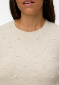 Pull léger beige avec une finition texturée et de petits points régulièrement espacés. Il a un col côtelé et est fabriqué dans un matériau tricoté doux.