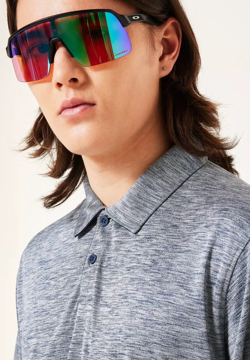 Joven con cabello oscuro de longitud media que lleva una camisa polo gris con botones y grandes gafas de sol deportivas reflectantes.