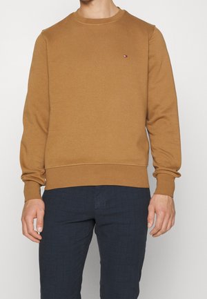 Mand iført en kamelbrun rundhalset sweatshirt med et lille rødt, hvidt og blåt logo på venstre bryst og mørkeblå bukser.