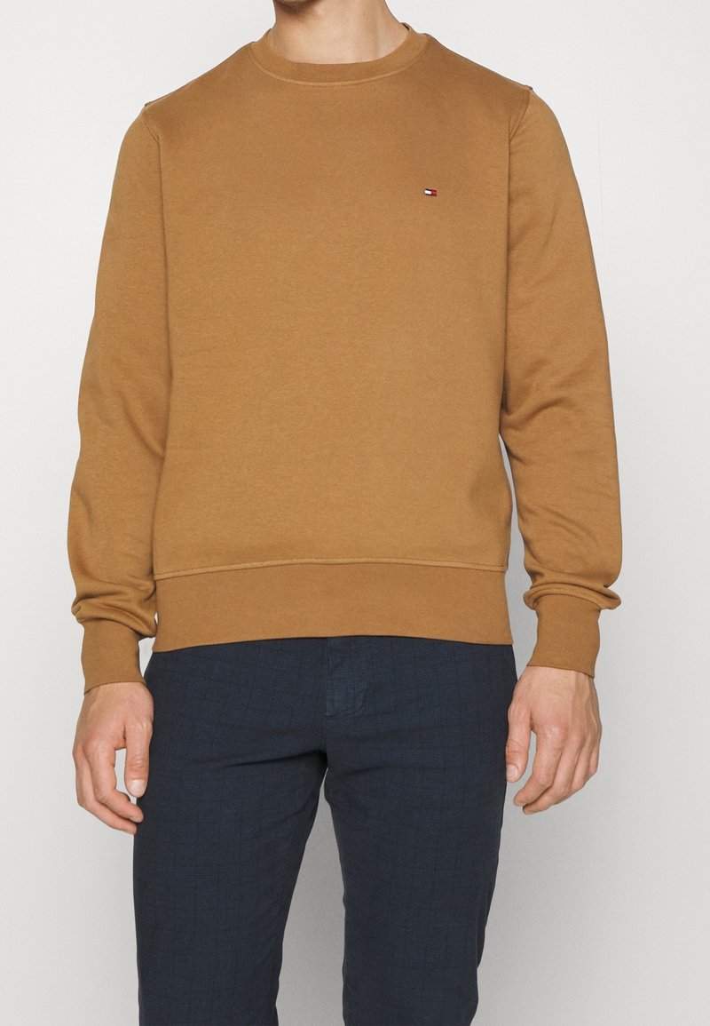 Homme portant un sweat-shirt à col rond couleur camel avec un petit logo rouge, blanc et bleu sur la poitrine gauche et un pantalon bleu foncé.
