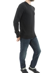 Czarny sweter z długim rękawem i okrągłym dekoltem, wykonany z dzianiny. Połączony z niebieskimi dżinsami i ciemnym obuwiem, stoi z rękami w kieszeniach.