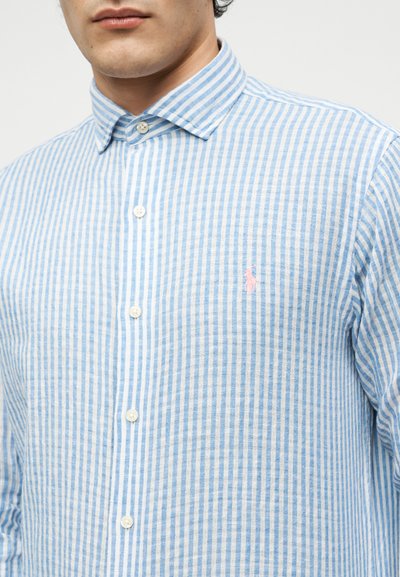 Άνδρας που φοράει μπλε και λευκό κάθετα ριγέ πουκάμισο με κουμπιά, γιακά και ένα μικρό ροζ κεντημένο λογότυπο στο στήθος.