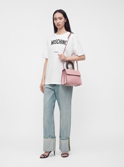 Jeune femme debout portant un t-shirt blanc Moschino, un jean clair retroussé, des sandales noires à talons et tenant un sac à main rose avec une poignée courbée.