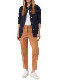 Giacca di jeans blu scuro con cuciture chiare, top bianco, pantaloni marrone-arancio e sneakers bianche. Outfit casual a strati.