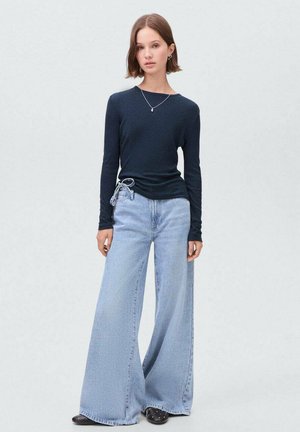 Laivansinis pitkähihaine paita, jossa on teksturoitu kangas, yhdistettynä korkeavyötäröisiin vaaleansinihin leveälahkeisiin farkkuihin. Mustat kengät täydentävät asun.