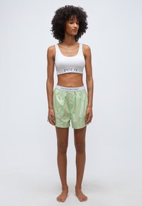 Γυναίκα όρθια, ξυπόλητη, με λευκό crop top με τη λέξη "POLO" και πράσινα ριγέ σορτς με ζωνάρι μέσης "POLO RALPH LAUREN" σε απλό φόντο.