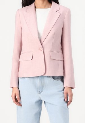 Frau trägt einen hellrosa Blazer mit einem Knopf, Pattentaschen, weißem Top und hellblauen Jeans.