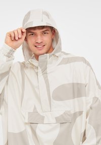 Vit vattentät jacka med stora grå abstrakta mönster, utrustad med huva, dragsnören och en framficka. Slätt, lätt material.