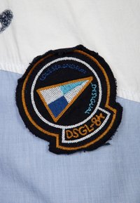 Gestickter Patch mit einem blauen und orangen dreieckigen Design auf schwarzem Hintergrund, mit weißen Akzenten und dem Text "DSGL8K", angebracht auf hellblauem Stoff.