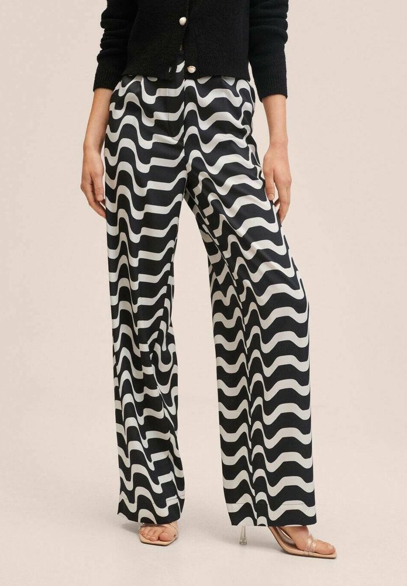 Personne portant un pantalon large à motif ondulé noir et blanc, un cardigan noir boutonné et des sandales à talons transparents, debout sur un fond beige.