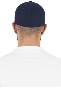 Cappellino da baseball blu navy con visiera piatta, realizzato in tessuto testurizzato. Regolatore sul retro. Indossato con una polo bianca, che mostra un colletto pulito.