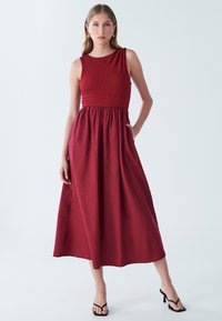 Crimson maxi obleka z raztegljivim bodijem, brezrokavnim dizajnom in stranskimi žepi. Iz lahkega blaga z dihajočo silhueto.