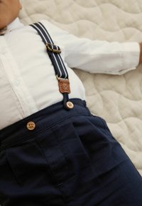 Temno modre naramnice z belimi črtastimi poudarki, pripete na svetlo obarvane hlače z lesenimi gumbi in teksturirano belo majico.