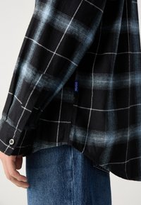 Gros plan d'une personne portant une chemise à carreaux noire et bleue avec une étiquette Hugo et un jean en denim bleu, montrant les détails sur le côté et les manches.