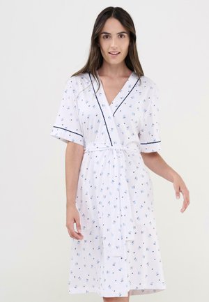 Mujer vistiendo un vestido blanco hasta la rodilla con estampados florales azules, mangas cortas, borde negro y un cinturón anudado en la cintura sobre un fondo liso.