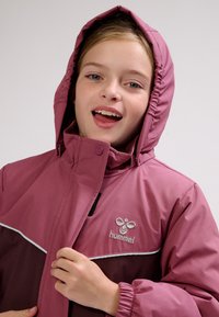 Pige smilende med åben mund iført en mørkerød og mørkelilla hættetrøje, der holder lynlåsen nær brystet med den ene hånd.