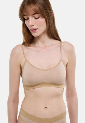 Femme à la peau claire et aux cheveux bruns portant un bralette à rayures beige et blanc et des sous-vêtements assortis, regardant vers le bas sur un fond uni.