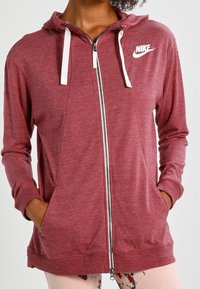 Red zip-up hoodie made of textured fabric, featuring a white Nike logo, drawstrings, and front pockets.
