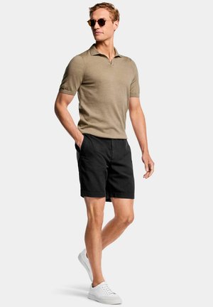 Uomo che indossa una polo beige a maniche corte, pantaloncini neri, scarpe da ginnastica bianche e occhiali da sole, in piedi con una mano in tasca su uno sfondo semplice.