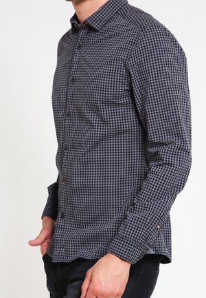 Mann trägt ein langärmeliges, geknöpftes, dunkelgrau-schwarz kariertes Hemd mit Kragen und steht vor einfachem Hintergrund.