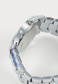 Reloj de pulsera de metal plateado con una caja lisa y brillante, un bisel texturizado y una pulsera con Acabados en cristal azul.