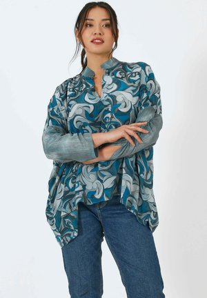 Blouse à manches longues avec un col mandarin, présentant un motif tourbillonnant bleu et gris sur un tissu léger, associée à un jean en denim.