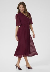 Burgundy pleated dress with short sheer sleeves and a v-neckline, featuring a fitted waist and flowing skirt. Paired with matching heeled shoes.