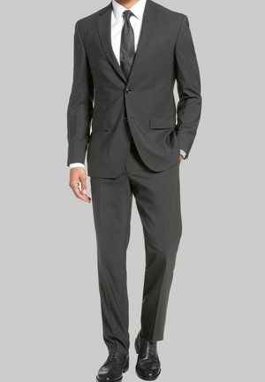 Costume gris ajusté avec revers cranté, fermeture à deux boutons et pantalon à pli plat. Associe à une chemise blanche et une cravate noire, terminé par des chaussures noires.