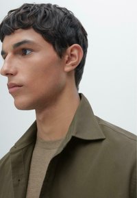 Olijfgroene jas met een zachte textuur en puntige kraag, gecombineerd met een neutraal beige shirt. Close-up profielweergave van het model.