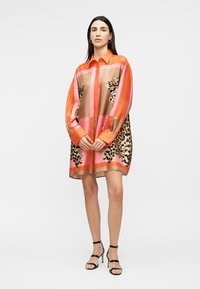 Γυναίκα φοράει φόρεμα με μακριά μανίκια και κουμπιά, με πορτοκαλί, ροζ και λεοπάρ πάνελ, συνδυασμένο με μαύρα πέδιλα με ψηλό τακούνι.