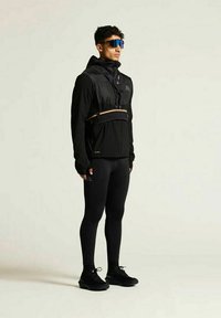 Schwarze sportliche Jacke mit Kapuze, strukturiertem Gewebe, Brustgurten, getragen über figurbetonten schwarzen Leggings und leichten schwarzen Sneakers. Blaue Sonnenbrille.