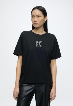 Otse mustade juustega naine kannab musta Karl Lagerfeld logoga t-särki ja musti nahkpükse, seisab valgel ühevärvilisel taustal.