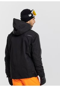 Giacca nera isolata con cappuccio, polsini elasticizzati e orlo con coulisse. Indossata con pantaloni da neve arancioni e berretto nero, in abbinamento a occhiali da sci.