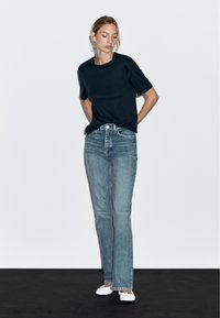 T-shirt noir à manches courtes associé à un jean flare bleu clair et des chaussures blanches à enfiler, sur un fond uni.