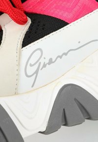 Section d'une basket multicolore montrant des lacets rouges, un mesh noir, du cuir blanc avec signature, et une semelle texturée grise et blanche.