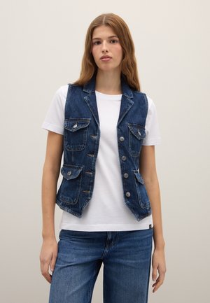 Jeune femme debout, portant un t-shirt blanc, un gilet en denim bleu avec des boutons et des poches, et un jean bleu sur un fond uni.