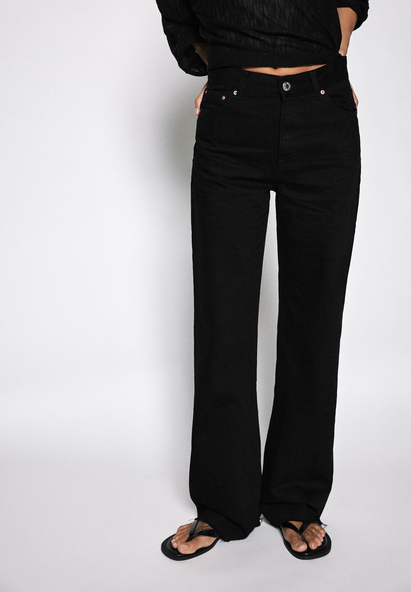 Personne portant un pantalon noir taille haute à jambes larges et des tongs noires, debout les mains dans les poches devant un fond uni.