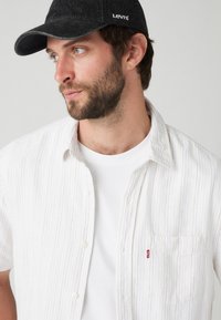 Camicia bianca a maniche corte con righe verticali multicolori, con bottoni, dotata di tasca sul petto sinistro e un'etichetta rossa Levi's. Cappellino nero con logo.