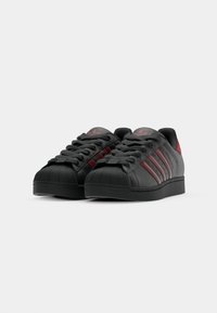 Čierne kožené tenisky s červenými akcentmi, s klasickým designom shell toe, šnúrkami a trojpruhovým brandingom na stranách.