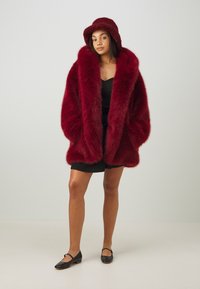 Rød faux fur-kåpe med stor krage og oversized passform, kombinert med en matchende rød bøttehatt. Modellen har på seg en svart kjole og leiligheter.